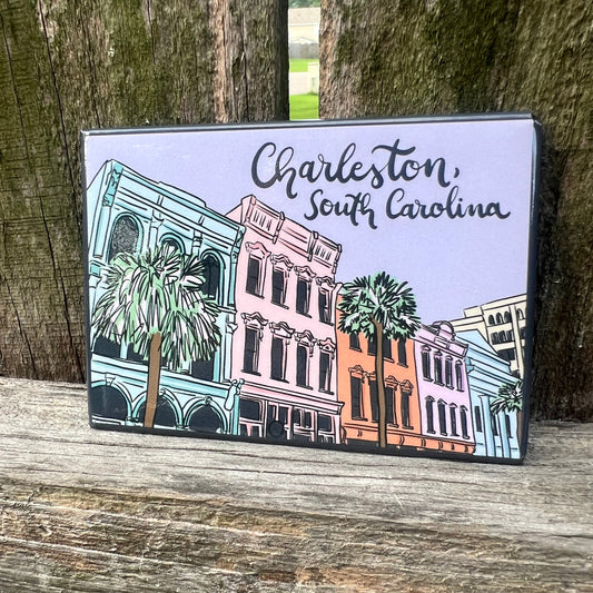 A magnet featuring a hand-drawn illustration of Charleston, South Carolina buildings and palm trees, placed on a weathered wooden surface in front of tree trunks.